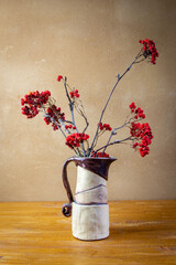 Autumn awesome still life with Sorbus aucuparia branches in handmade ceramic brown white vase placed on wooden board in front of brown wall with clay plaster