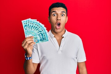 Young arab man holding yuan chinese banknotes scared and amazed with open mouth for surprise, disbelief face