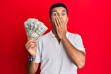 Young arab man holding dollars covering mouth with hand, shocked and afraid for mistake. surprised expression