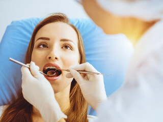 Smiling young woman with orthodontic brackets examined by dentist in sunny dental clinic. Healthy...