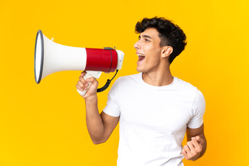 Young Argentinian man isolated on yellow background shouting through a megaphone to announce something in lateral position