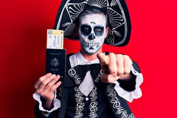 Young man wearing mexican day of the dead costume holding united states passport and boarding pass pointing with finger to the camera and to you, confident gesture looking serious