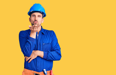 Young handsome man wearing worker uniform and hardhat looking at the camera blowing a kiss with hand on air being lovely and sexy. love expression.