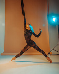 Fototapeta na wymiar young girl in black slim costume is standing on the white floor in gymnastic pose on the beige background in photo studio in warm and cold lights, sport concept, free space