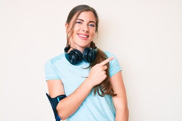 Fototapeta premium Beautiful young caucasian woman wearing gym clothes and using headphones cheerful with a smile of face pointing with hand and finger up to the side with happy and natural expression on face