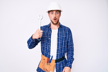 Young handsome man weaing handyman uniform holding wrench scared and amazed with open mouth for surprise, disbelief face