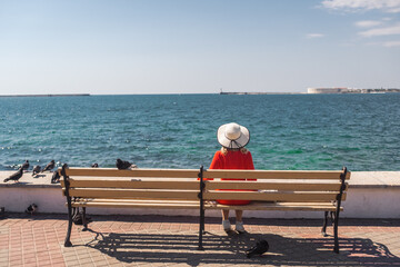 a girl in a red dress and hat sits on a bench in front of the sea and looks at it