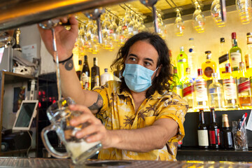 Front view of a waiter pouring a pitcher of beer in a bar. He is wearing a medical mask.