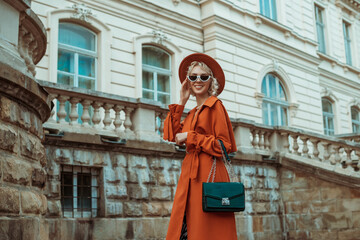 Happy smiling fashionable woman wearing orange trench coat, hat, sunglasses holding green faux reptile textured bag, posing in street of European city. Copy, empty space for text