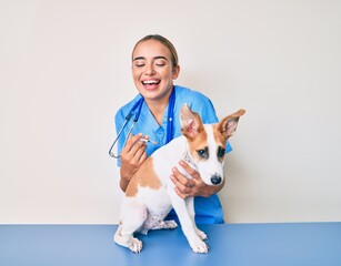 Young beautiful blonde veterinarian woman putting vaccine to puppy dog smiling and laughing hard out loud because funny crazy joke.