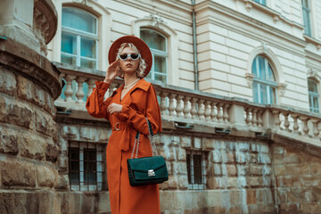 Outdoor autumn portrait of elegant woman wearing long orange trench coat, hat, sunglasses, with green croco textured bag, posing in street of European city. Copy, empty space for text