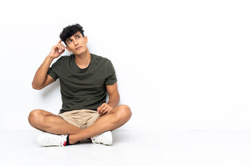 Young Argentinian man sitting on the floor having doubts and with confuse face expression