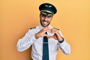 Handsome hispanic man wearing airplane pilot uniform smiling in love doing heart symbol shape with hands. romantic concept.
