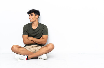 Young Argentinian man sitting on the floor with arms crossed and happy