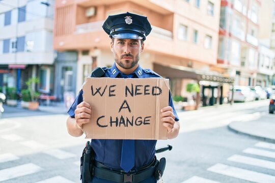 Young handsome hispanic policeman wearing police uniform with serious expression holding we need a change banner at town street