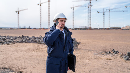 a civil engineer in a white helmet is talking on the phone in the background of construction.