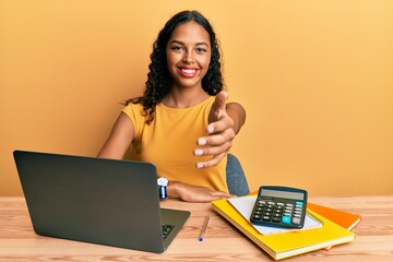Young african american girl working at the office with laptop and calculator smiling friendly offering handshake as greeting and welcoming. successful business. - obrazy, fototapety, plakaty
