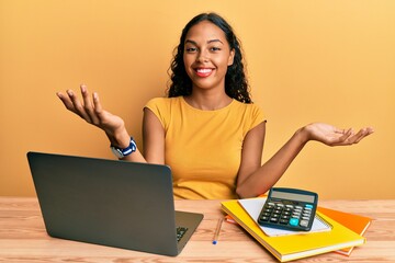Young african american girl working at the office with laptop and calculator smiling cheerful with open arms as friendly welcome, positive and confident greetings - obrazy, fototapety, plakaty