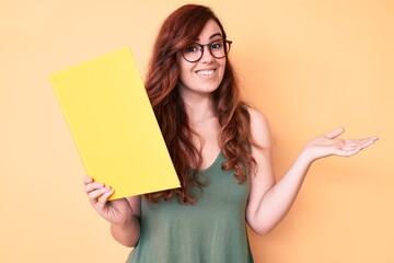 Naklejka premium Young beautiful woman wearing glasses holding book celebrating achievement with happy smile and winner expression with raised hand