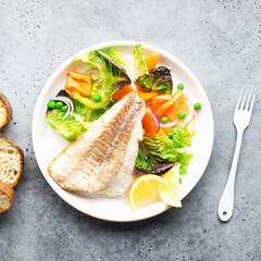 Fried fillet of sea white cod fish with juicy lettuce, capsicum, lemon, green peas on a large white dish on a gray background. Healthy balanced food. Top view,
