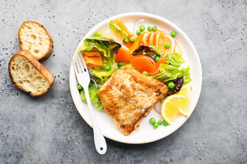 Fried fillet of sea white cod fish with juicy lettuce, capsicum, lemon, green peas on a large white dish on a gray background. Healthy balanced food. Top view, - obrazy, fototapety, plakaty