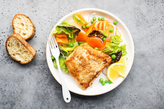 Fried fillet of sea white cod fish with juicy lettuce, capsicum, lemon, green peas on a large white dish on a gray background. Healthy balanced food. Top view,