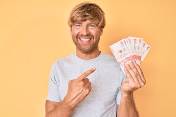 Young blond man holding colombian pesos smiling happy pointing with hand and finger