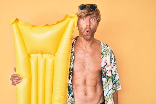 Young blond man wearing summer style and holding matress float scared and amazed with open mouth for surprise, disbelief face