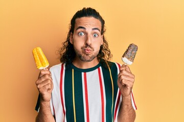 Young handsome man with long hair eating two ice cream puffing cheeks with funny face. mouth inflated with air, catching air. - obrazy, fototapety, plakaty