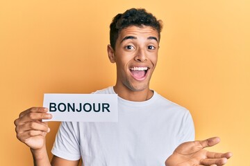 Young handsome african american man holding bonjour french greeting word celebrating achievement with happy smile and winner expression with raised hand © Krakenimages.com
