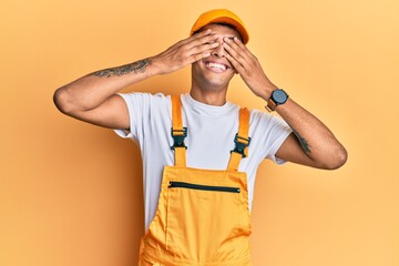 Young handsome african american man wearing handyman uniform over yellow background covering eyes with hands smiling cheerful and funny. blind concept.