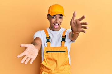 Young handsome african american man wearing handyman uniform over yellow background looking at the camera smiling with open arms for hug. cheerful expression embracing happiness.