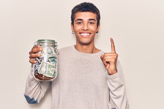 Young african amercian man holding jar with savings smiling with an idea or question pointing finger with happy face, number one
