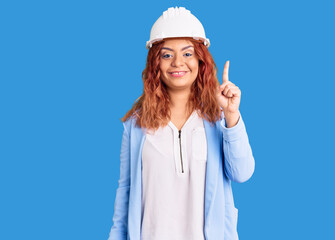 Young latin woman wearing architect hardhat showing and pointing up with finger number one while smiling confident and happy.