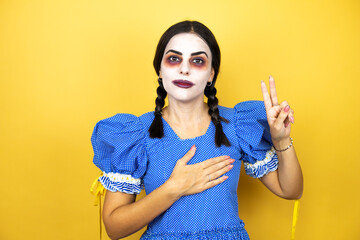 woman wearing a scary doll halloween costume over yellow background smiling swearing with hand on chest and fingers up, making a loyalty promise oath
