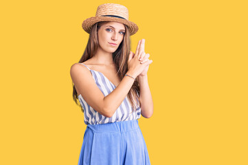 Fototapeta premium Young beautiful blonde woman wearing summer hat holding symbolic gun with hand gesture, playing killing shooting weapons, angry face
