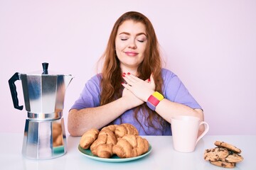 Young redhead woman eating breakfast very happy and excited doing winner gesture with arms raised, smiling and screaming for success. celebration concept.