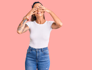 Young hispanic woman with tattoo wearing casual white tshirt covering eyes with hands smiling...