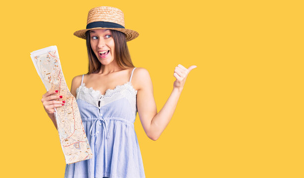 Beautiful brunette young woman wearing summer hat holding city map pointing thumb up to the side smiling happy with open mouth