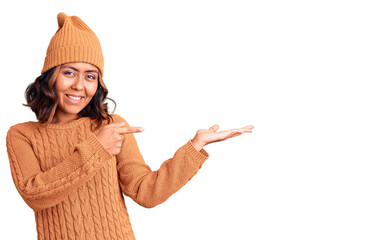 Young beautiful mixed race woman wearing wool sweater and winter hat amazed and smiling to the camera while presenting with hand and pointing with finger.