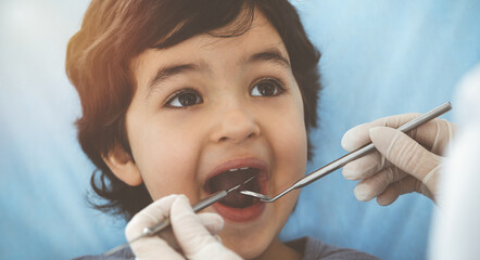 Cute arab boy sitting at dental chair with open mouth during oral checking up with doctor. Visiting sunny dentist office