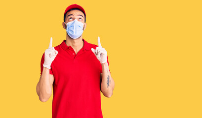 Young handsome hispanic man wearing delivery uniform and medical mask amazed and surprised looking up and pointing with fingers and raised arms.