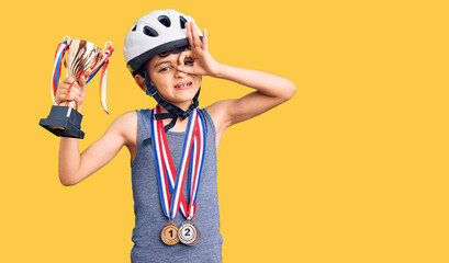 Little cute boy kid wearing bike helmet and winner medals holding winner trophy smiling happy doing ok sign with hand on eye looking through fingers