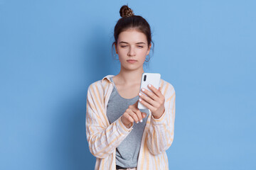 Brunette lady has combed hair, holds modern cell phone, wears casual shirt isolated over blue background, brunette female typing messages, looks at device's screen.