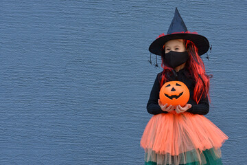A little girl in a witch costume in a medical mask holding a bucket of pumpkin for sweets on...
