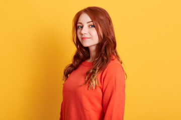 Fototapeta premium Gorgeous red haired young female with confident look, poses for fashionable magazine, looks at camera, wears casual orange sweater, posing against yellow background.