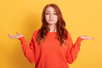 Confusion very offended red haired woman looking at camera with sad look and spreading her hands, wearing casual clothing, posing isolated over yellow wall.