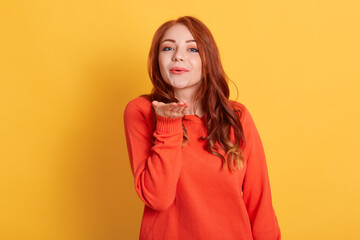 Portrait of pretty friendly girl in casual orange jumper, blowing air kiss with pout lips palm at camera isolated on yellow background, red haired saying goodbye to somebody.