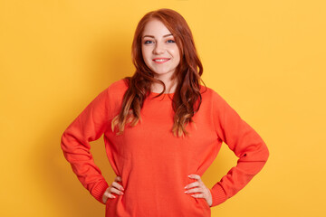 Confident young lady with red hair standing with hands on hips and looking at camera, being in good mood, smiling happily against yellow background, female wearing casual attire.