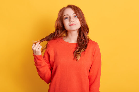 Unimpressed Indifferent Attractive Woman With Red Hair, Pulling Her Curls, Feeling Bored, Being On Boring Date, Standing Annoyed And Tired Over Yellow Background, Wearing Orange Jumper.